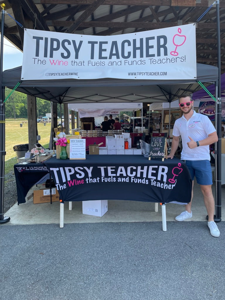 Person standing behind a 'Tipsy Teacher' booth at an outdoor event.